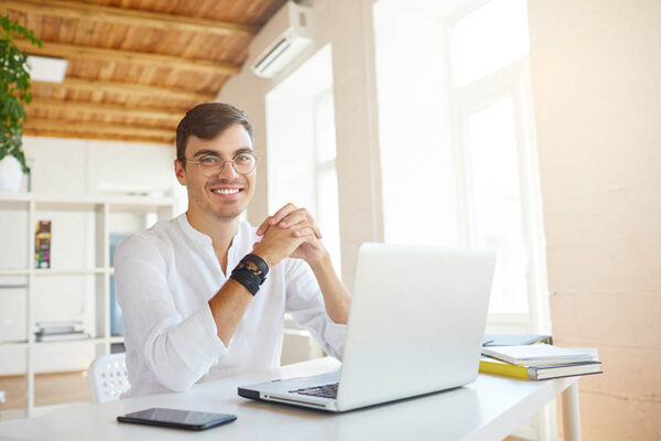 Portrait of happy successful young businessman wears white shirt and spectacles using laptop and cell phone sitting at the table in office and smiling
